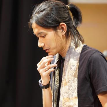 Jay Afrisando, a thirty-something Asian man composer/sound artist, with his right index and thumb on his chin as he pauses his speech during a concert opening.